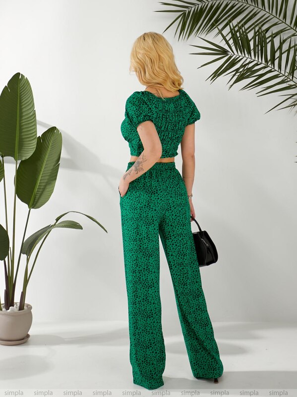 Woman in a green patterned outfit standing in front of a white wall with plants.