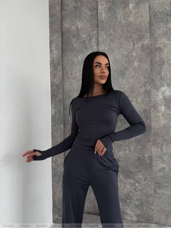 Woman wearing a dark gray long-sleeve top and pants against a gray stone wall.