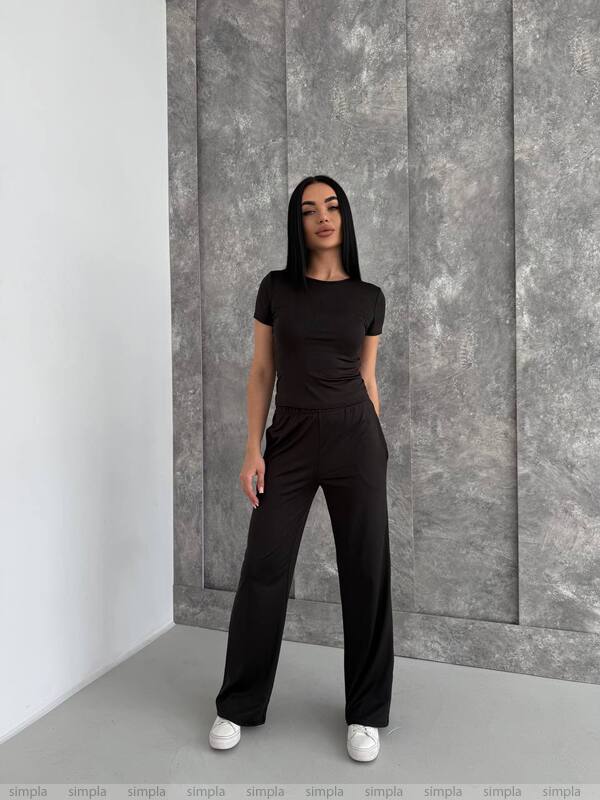 Woman wearing a black outfit standing against a gray paneled wall.