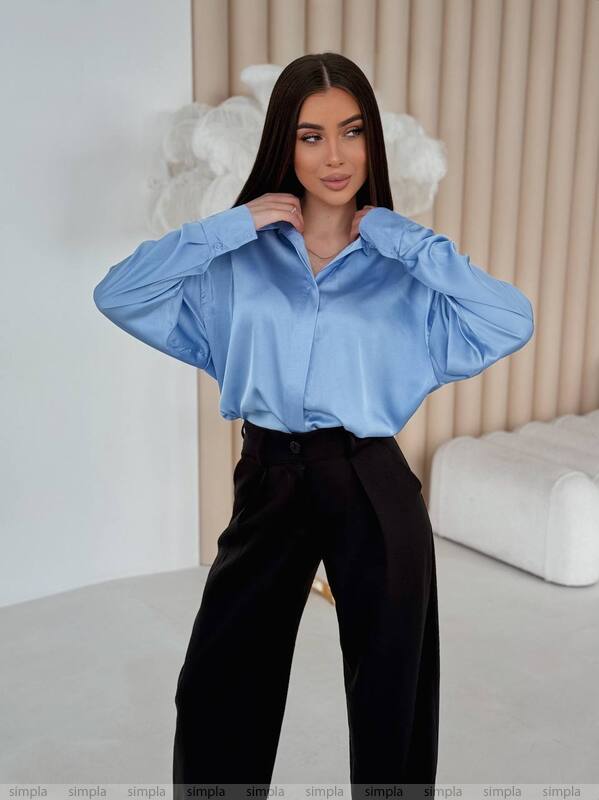Woman wearing a light blue blouse and black pants in a minimalistic room.