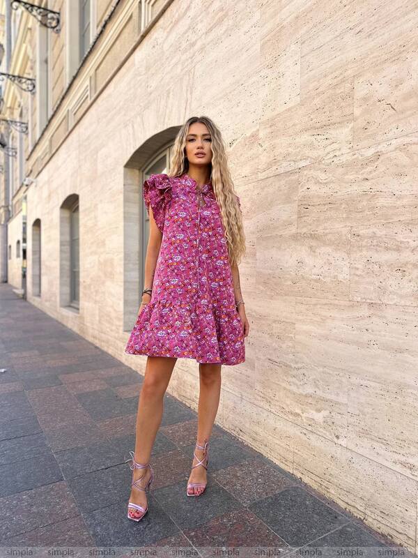 Woman in a pink floral dress standing against a beige wall on a city street.