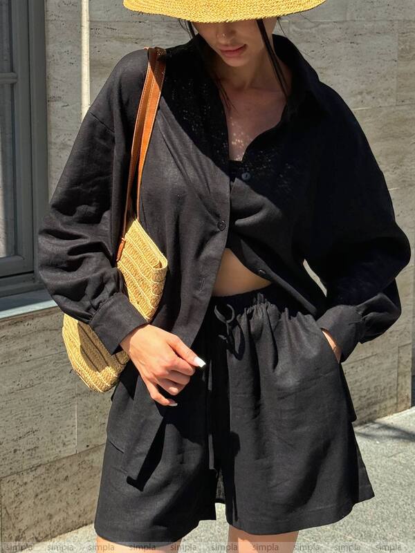Person wearing a black outfit with a straw hat and bag, standing against a stone wall.