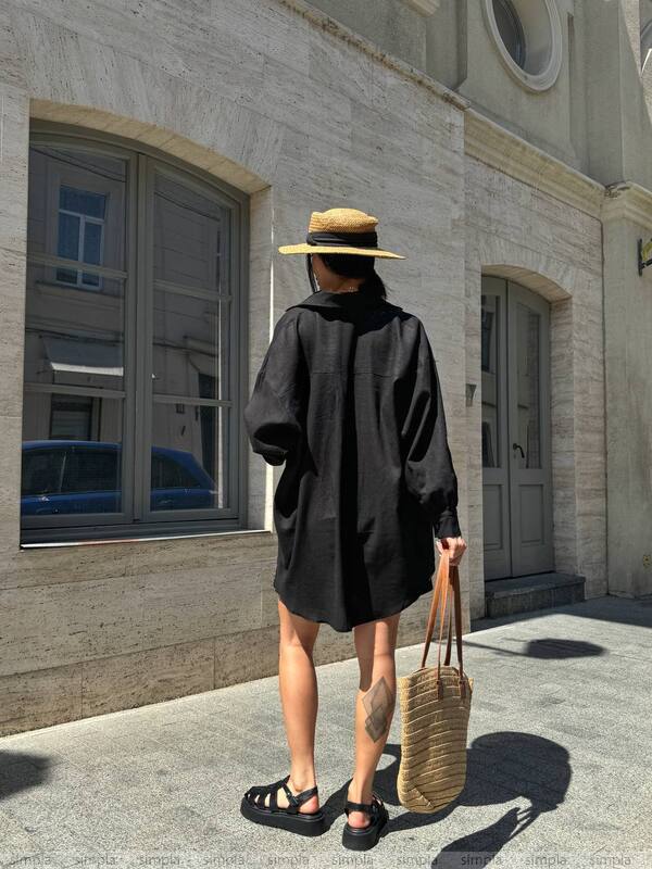 Person wearing a black coat and straw hat, holding a woven bag, standing in front of a building.