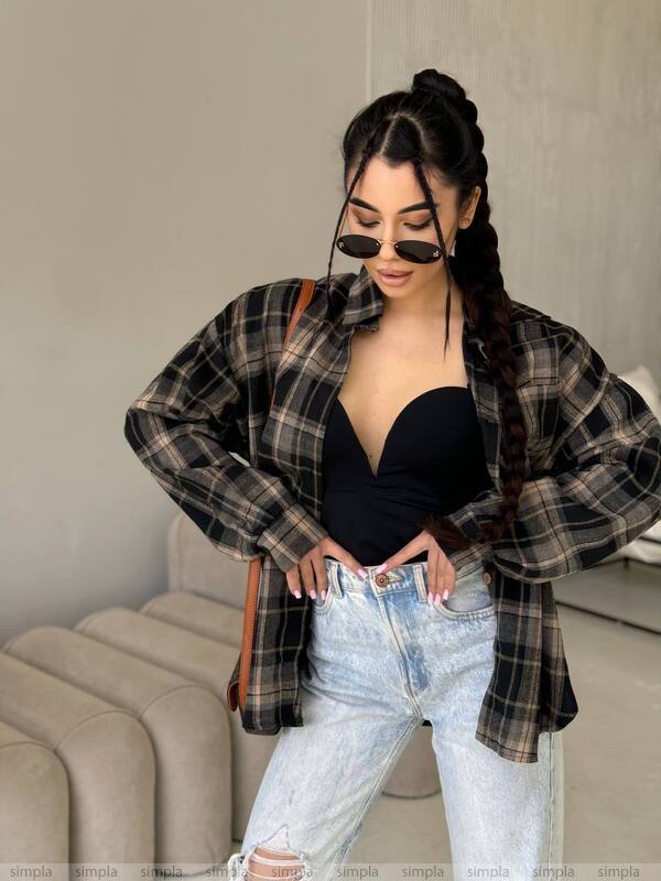 Woman wearing a plaid shirt over a black top and light blue jeans, standing in a room.