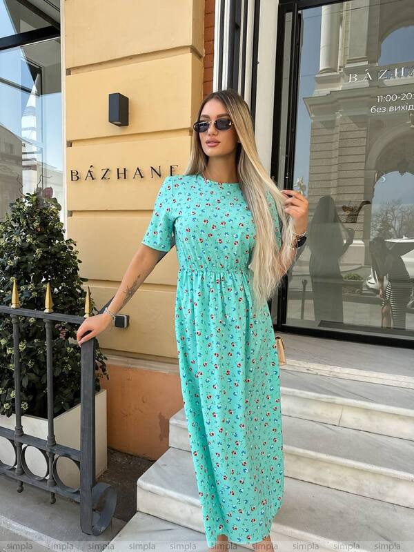 Woman in a turquoise dress standing outside a building with 'Bázhane' on the wall.