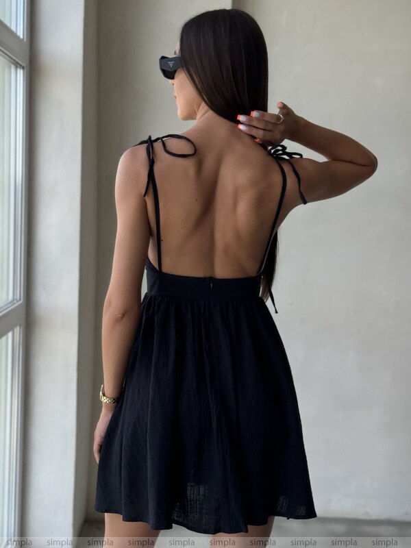 Woman wearing a black dress with thin straps, standing in a room with a window.