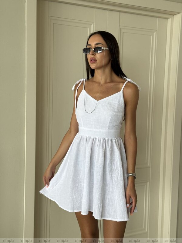 Woman wearing a white dress standing against a neutral wall.