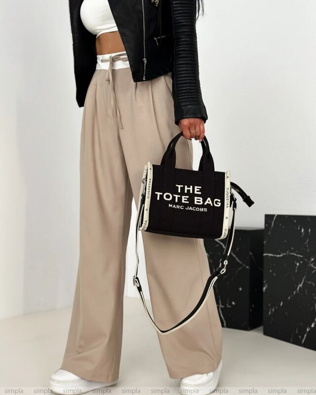 Person holding a black tote bag with 'The Tote Bag' text, wearing beige pants and a black jacket.