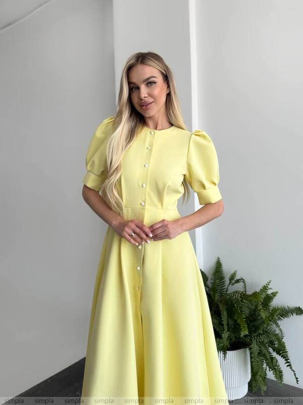 Woman wearing a yellow dress standing indoors with a plant in the background