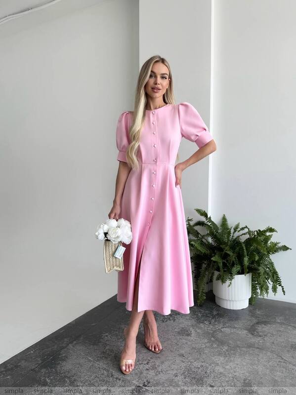 Woman wearing a pink dress standing indoors with a plant and white flowers in the background