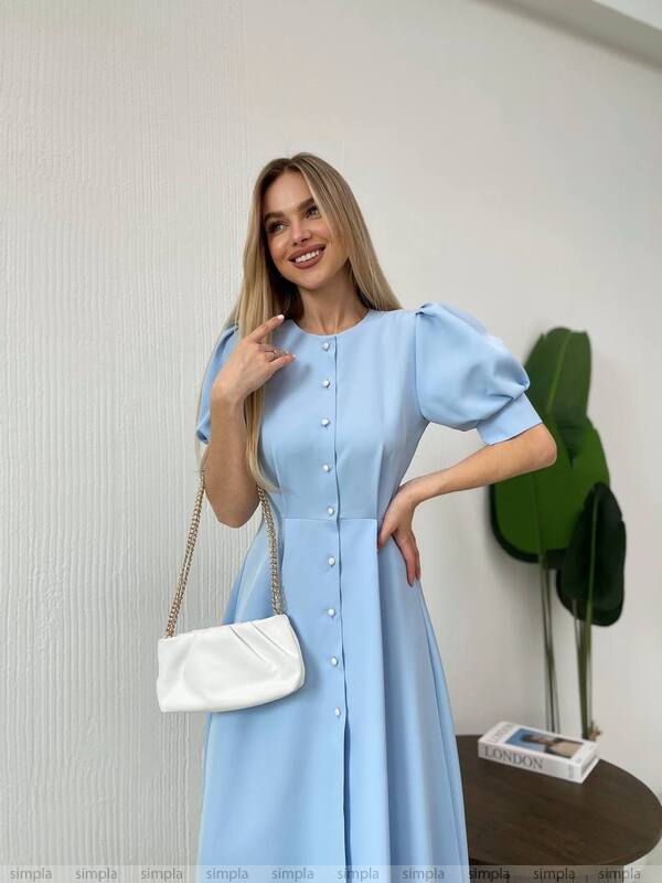 Woman wearing a light blue dress with a white handbag indoors.