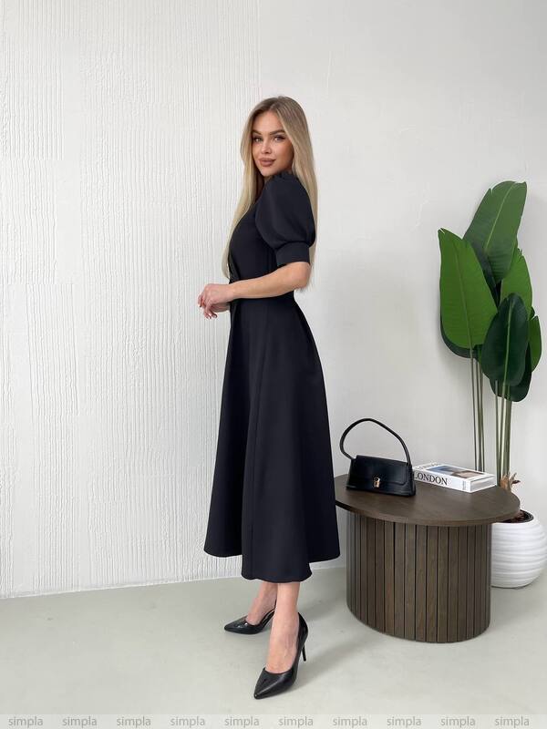 Woman in a black dress standing next to a wooden table with a plant and handbag.