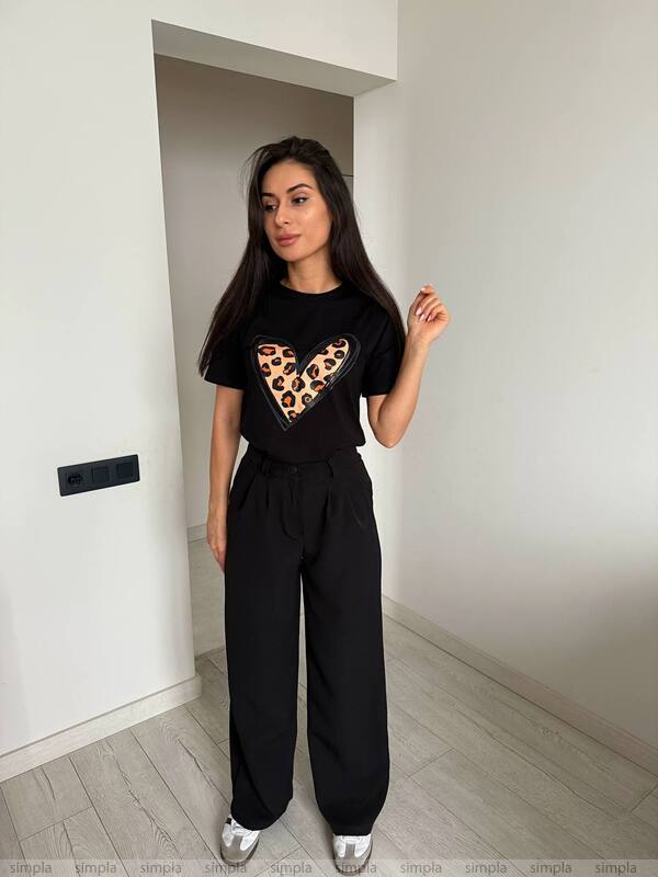 Woman wearing a black t-shirt with a leopard print heart design and black pants in a minimalistic room.