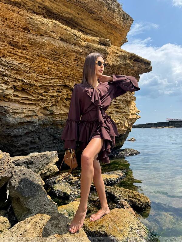 Woman in a brown dress standing on rocks by the water with a rocky cliff and blue sky in the background.