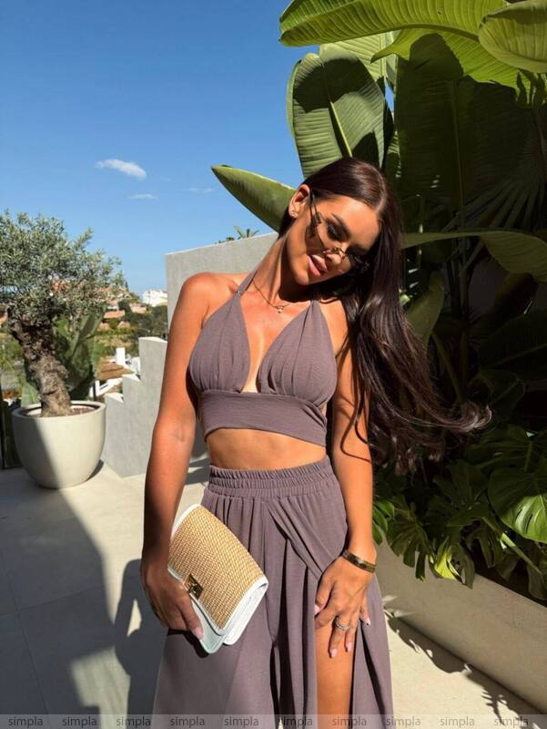 Woman in a brown outfit standing outdoors with plants and a clear sky in the background