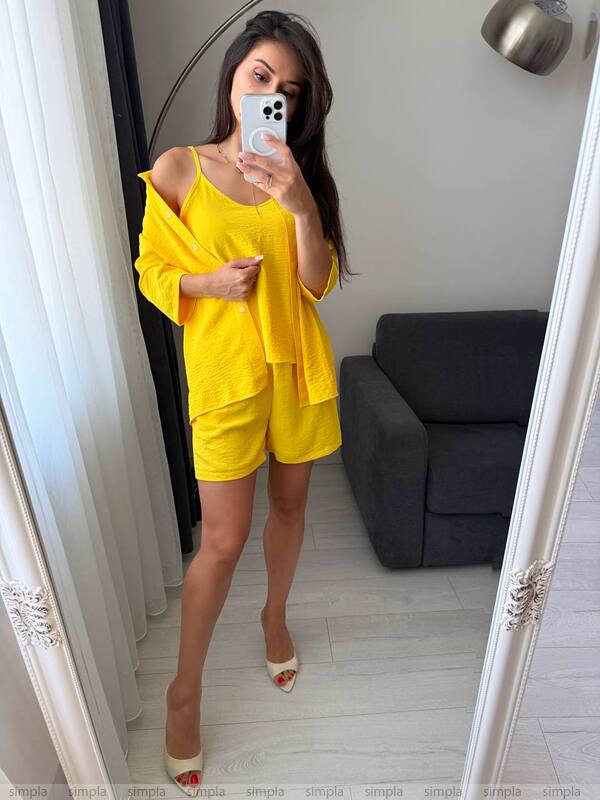 Woman taking a mirror selfie wearing a yellow outfit in a room with a gray sofa.