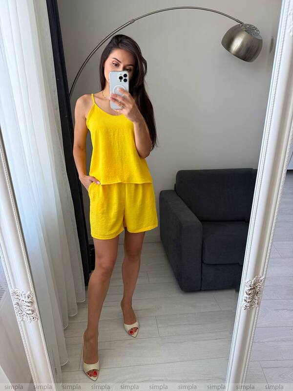 Woman taking a mirror selfie wearing a yellow outfit in a room with a gray sofa and white walls.