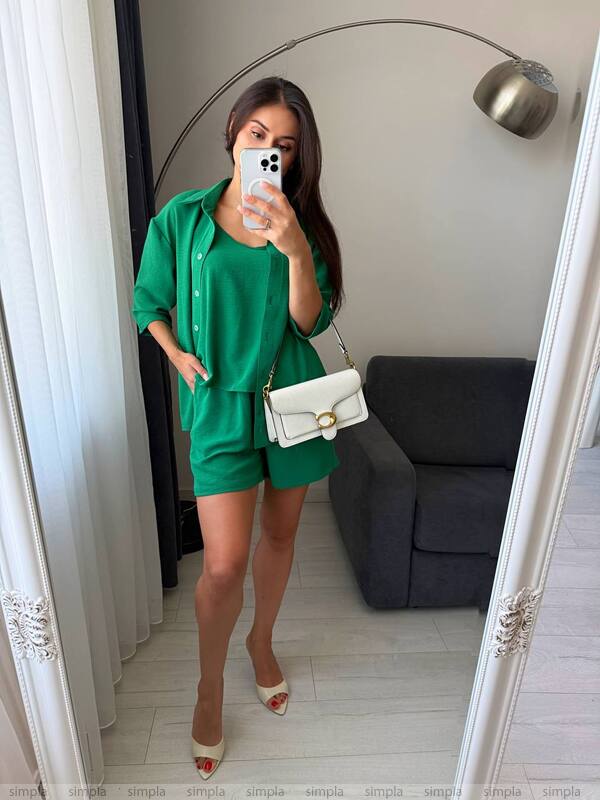 Woman taking a mirror selfie wearing a green outfit with a white handbag in a room.