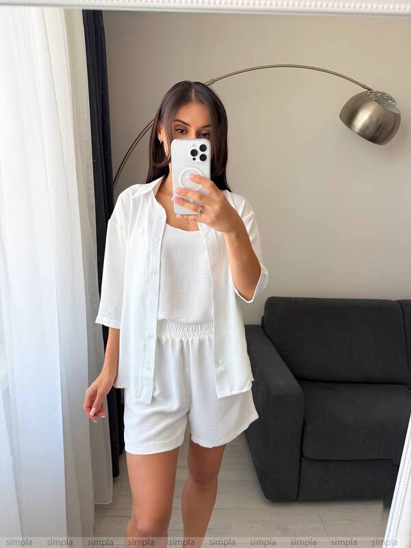 Woman taking a mirror selfie wearing a white outfit in a room with a gray sofa and white curtains.