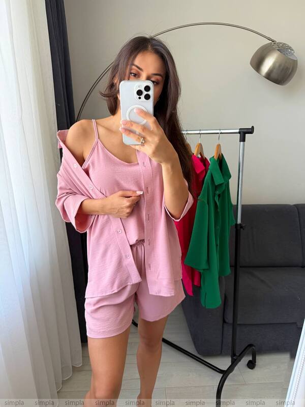 Person wearing a pink outfit taking a mirror selfie in a room with a coat rack and gray sofa.