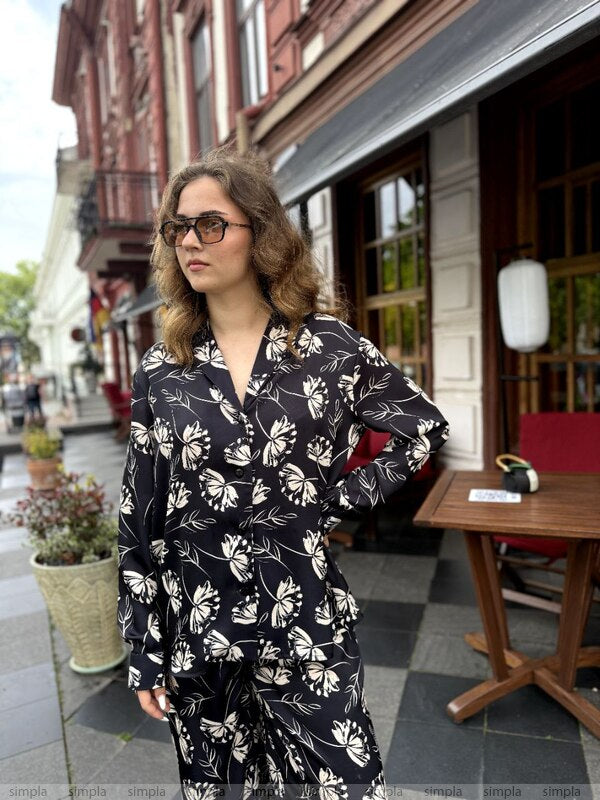 Woman wearing a black floral outfit standing on a street with buildings and tables in the background.