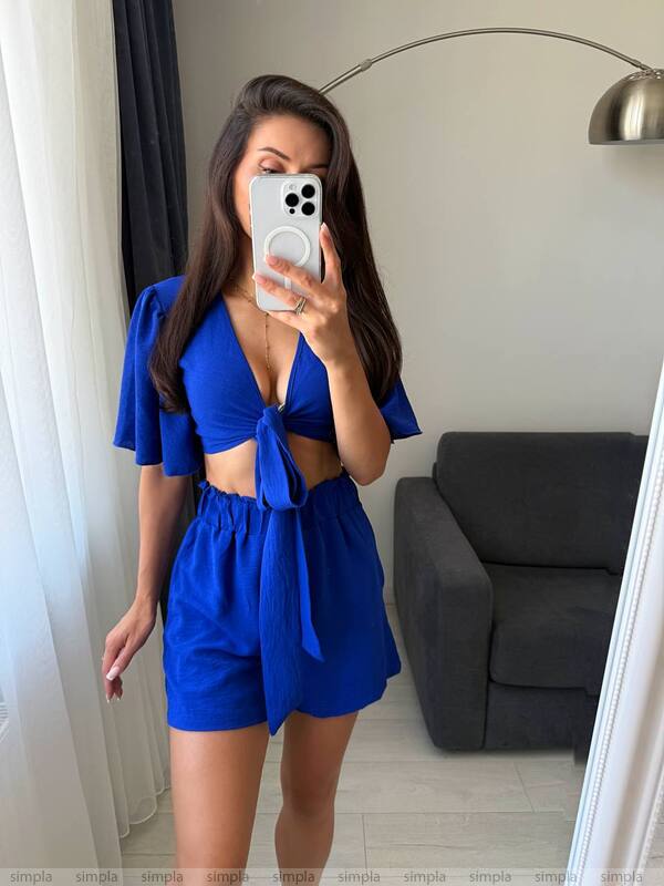 Woman taking a mirror selfie wearing a blue outfit in a room with a gray couch and white curtains.