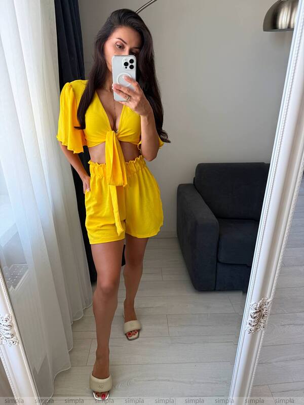 Woman taking a mirror selfie wearing a yellow outfit in a room with a gray sofa.