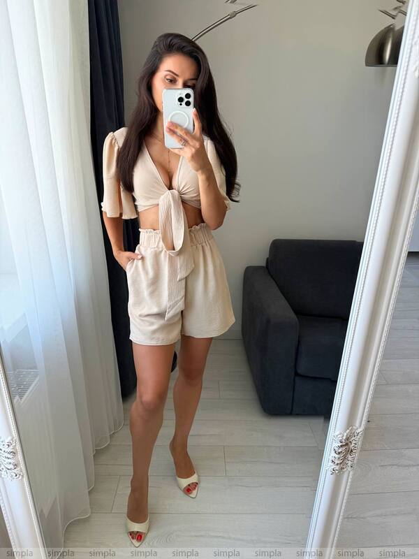 Woman taking a mirror selfie wearing a beige outfit in a room with a dark sofa.
