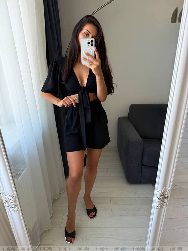 Woman taking a mirror selfie wearing a black outfit in a room with white curtains and a gray sofa.