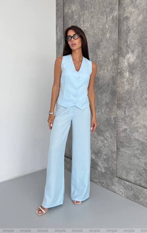 Woman wearing a light blue sleeveless top and wide-leg pants against a gray wall.