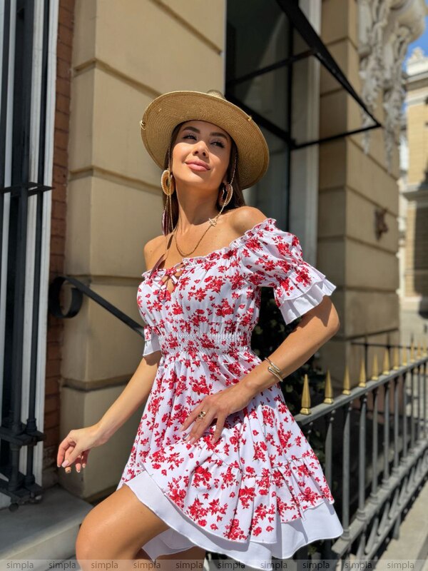Woman wearing a white dress with red floral patterns and a straw hat standing outdoors.
