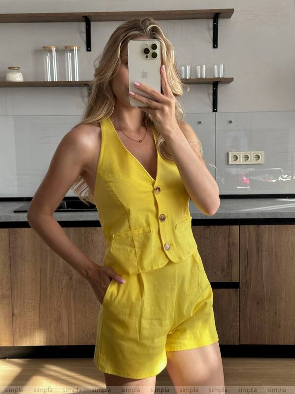 Woman taking a mirror selfie wearing a yellow romper in a kitchen.