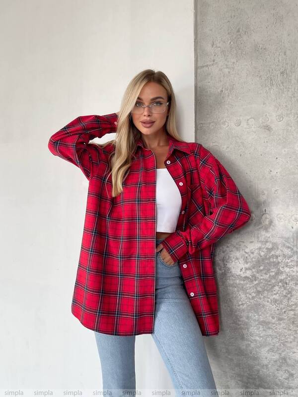 Woman wearing a red plaid shirt over a white top and light blue jeans against a gray wall.