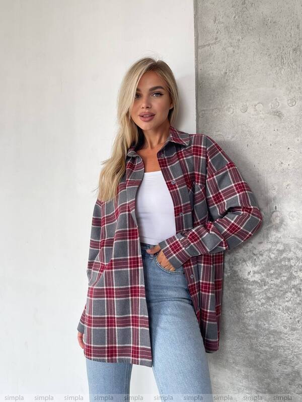 Woman wearing a plaid shirt over a white top and jeans against a concrete wall.
