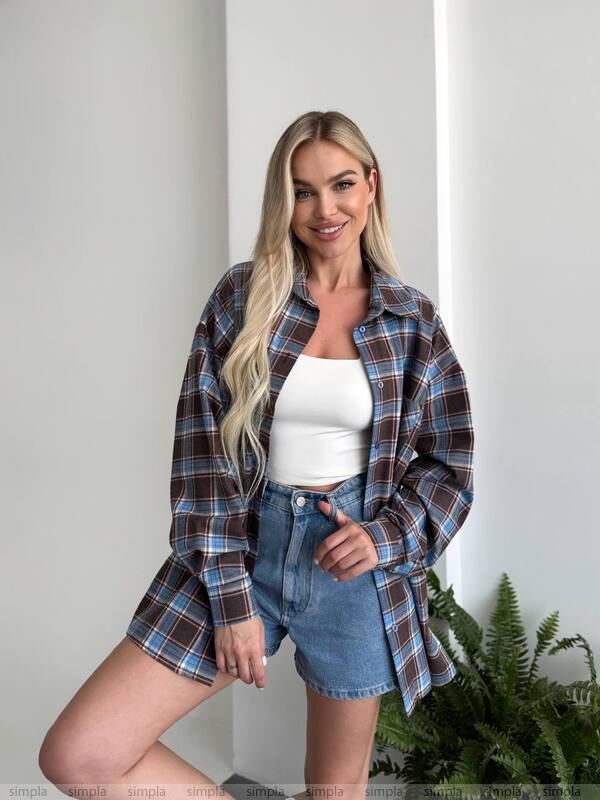 Woman wearing a plaid shirt over a white top and denim shorts, standing indoors with a plant in the background.