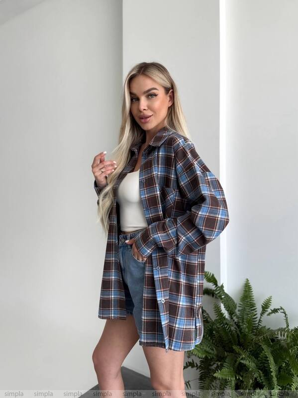 Woman wearing a plaid shirt over a white top and denim shorts, standing against a plain wall with a plant in the background.