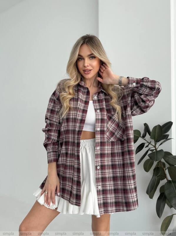 Woman wearing a plaid shirt over a white top and skirt, standing against a plain background with a plant.