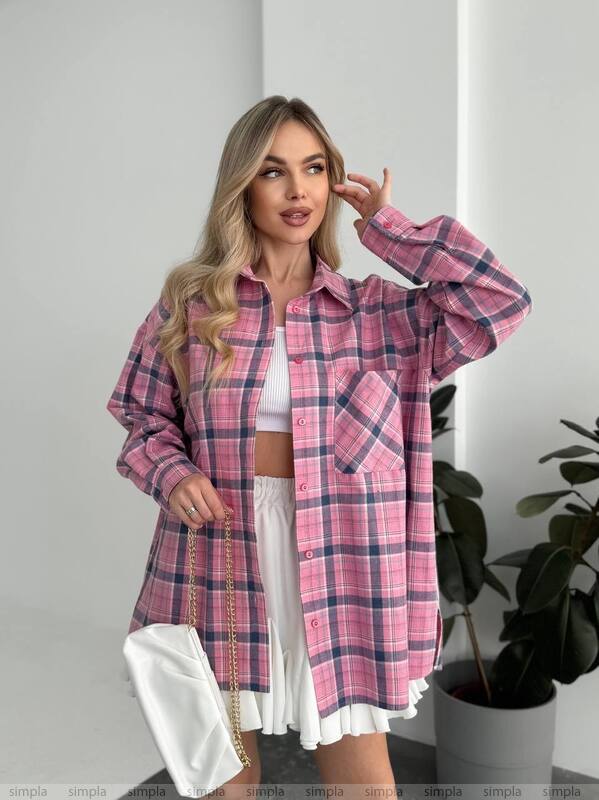 Woman wearing a pink plaid shirt over a white outfit, standing indoors with a plant in the background.