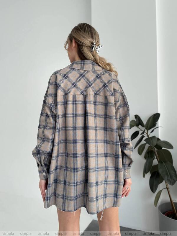 Person wearing a plaid shirt over a dress against a white wall with a plant on the right.