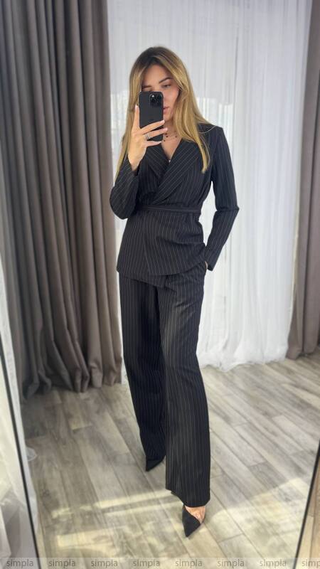 Woman taking a mirror selfie wearing a black pinstripe suit in a room with curtains.