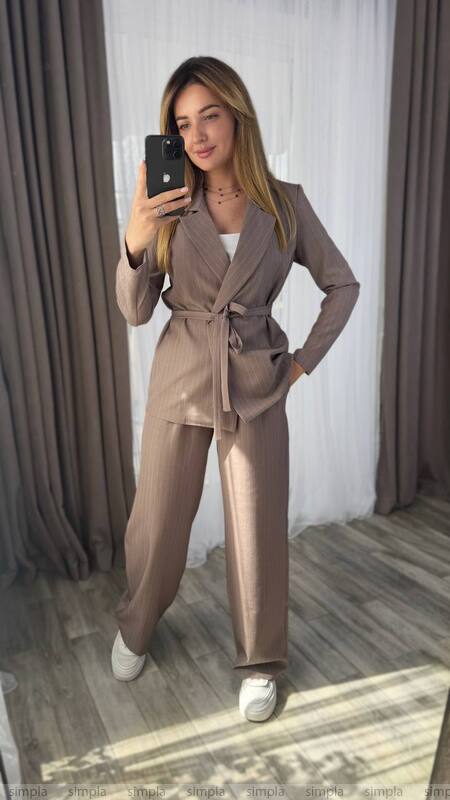 Woman taking a mirror selfie wearing a beige suit in a room with curtains.