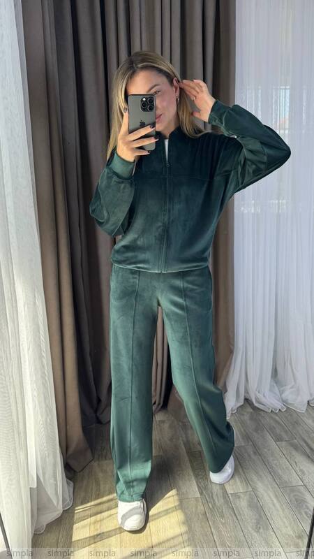 Person wearing a green tracksuit taking a mirror selfie in a room with white curtains.