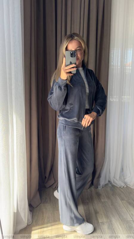 Person wearing a gray tracksuit taking a mirror selfie in a room with curtains.