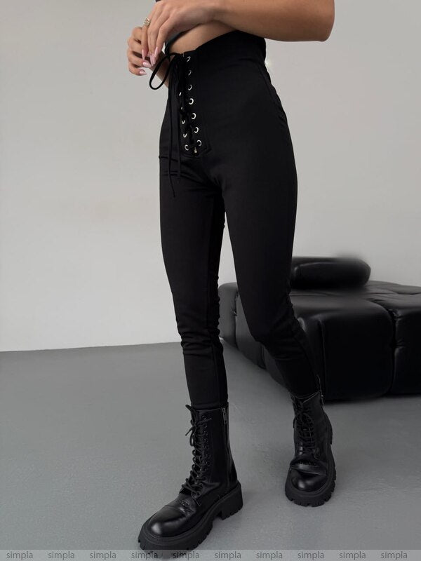 Person wearing black high-waisted pants with lace-up details and black boots on a gray floor.