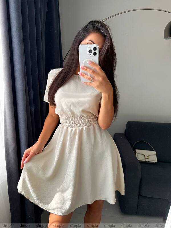 Woman taking a mirror selfie wearing a white dress in a room with a gray curtain and black sofa.