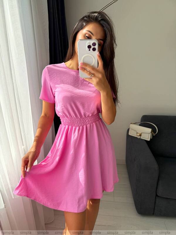 Woman wearing a pink dress taking a mirror selfie in a room with a couch and white curtains.