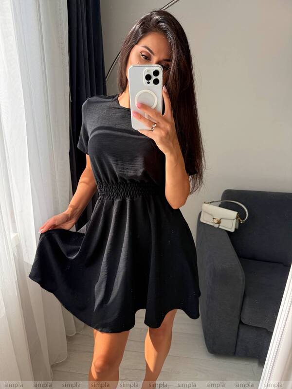 Woman wearing a black dress taking a mirror selfie in a room with a gray sofa.