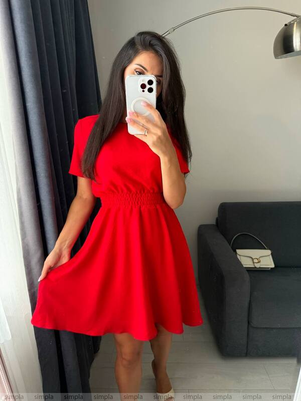 Woman taking a mirror selfie wearing a red dress in a room with a gray sofa and lamp.