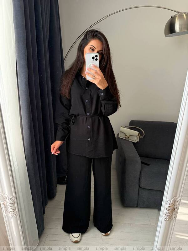 Person wearing a black outfit taking a mirror selfie in a room with a gray sofa and lamp.