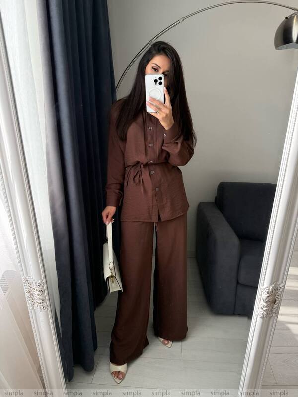 Person taking a mirror selfie wearing a brown outfit in a room with a gray sofa and black curtains.